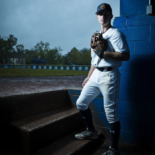 Baseball outfielder stands in dugout on a rainy day with the field behind him.