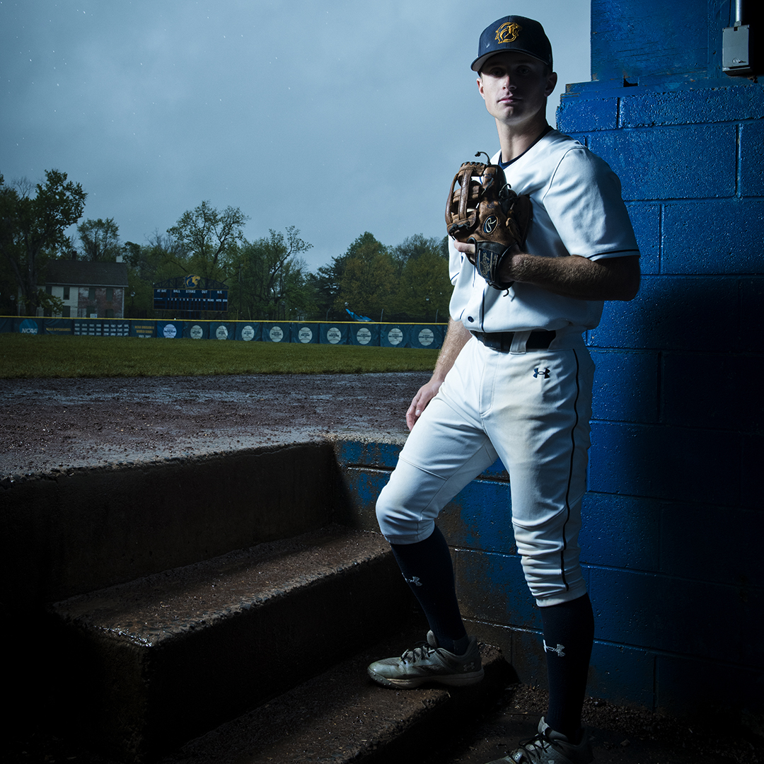 Baseball outfielder stands in dugout on a rainy day with the field behind him.